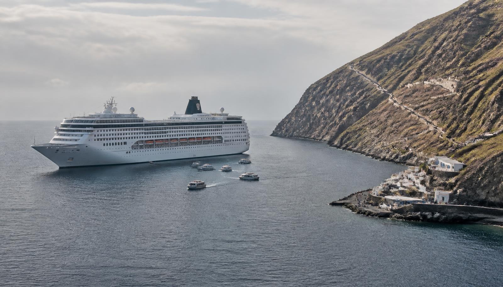 A cruise ship at anchor off a steep island coast with small tender boats running to a small harbour