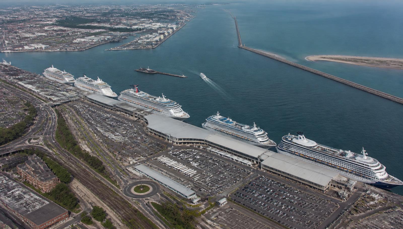 Aerial view of a cruise gateway port with long berths, terminal buildings, coach parking and a channel out to sea