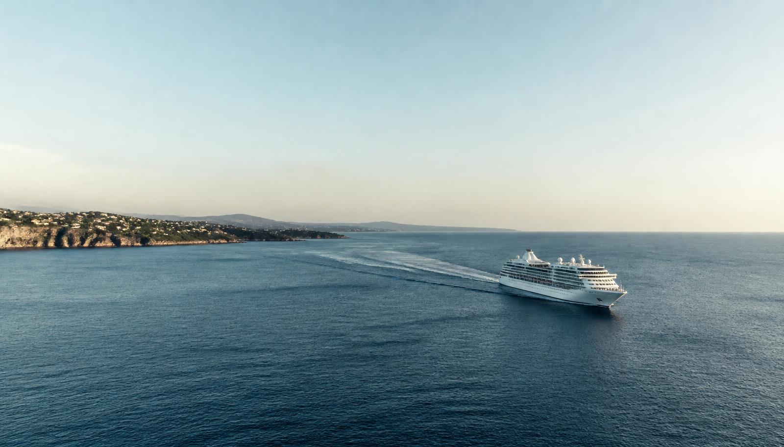 A cruise ship moving away from a coastline into open water in late afternoon light