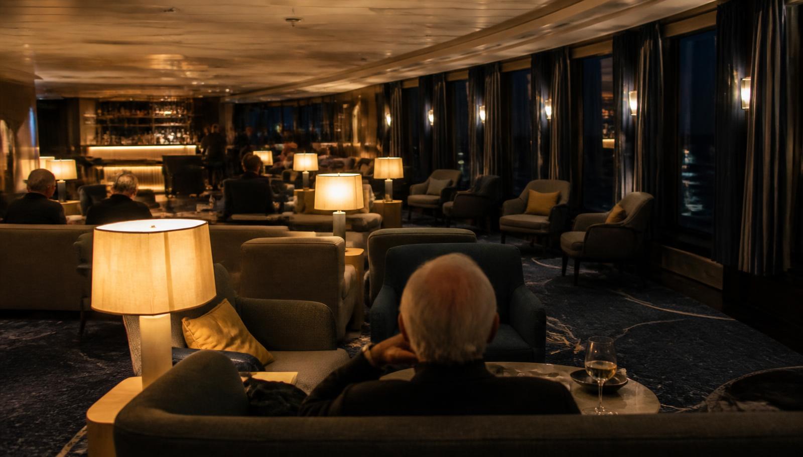 A ship lounge in the evening with low lamplight, seating groups and dark windows