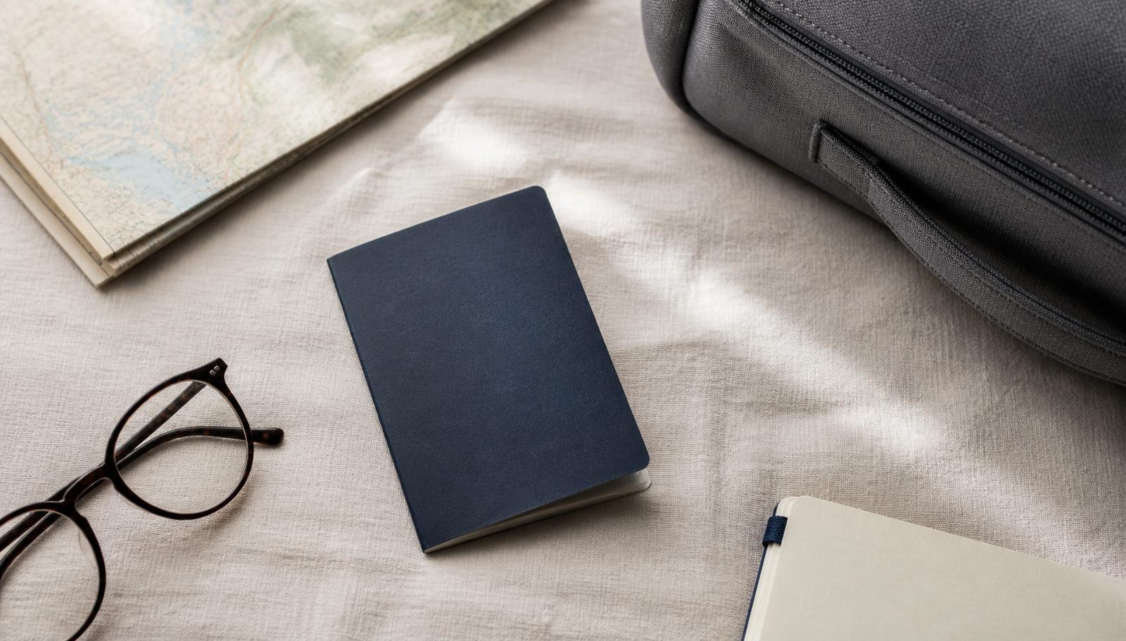 Travel documents, glasses, a notebook and a suitcase laid out on a linen surface in daylight
