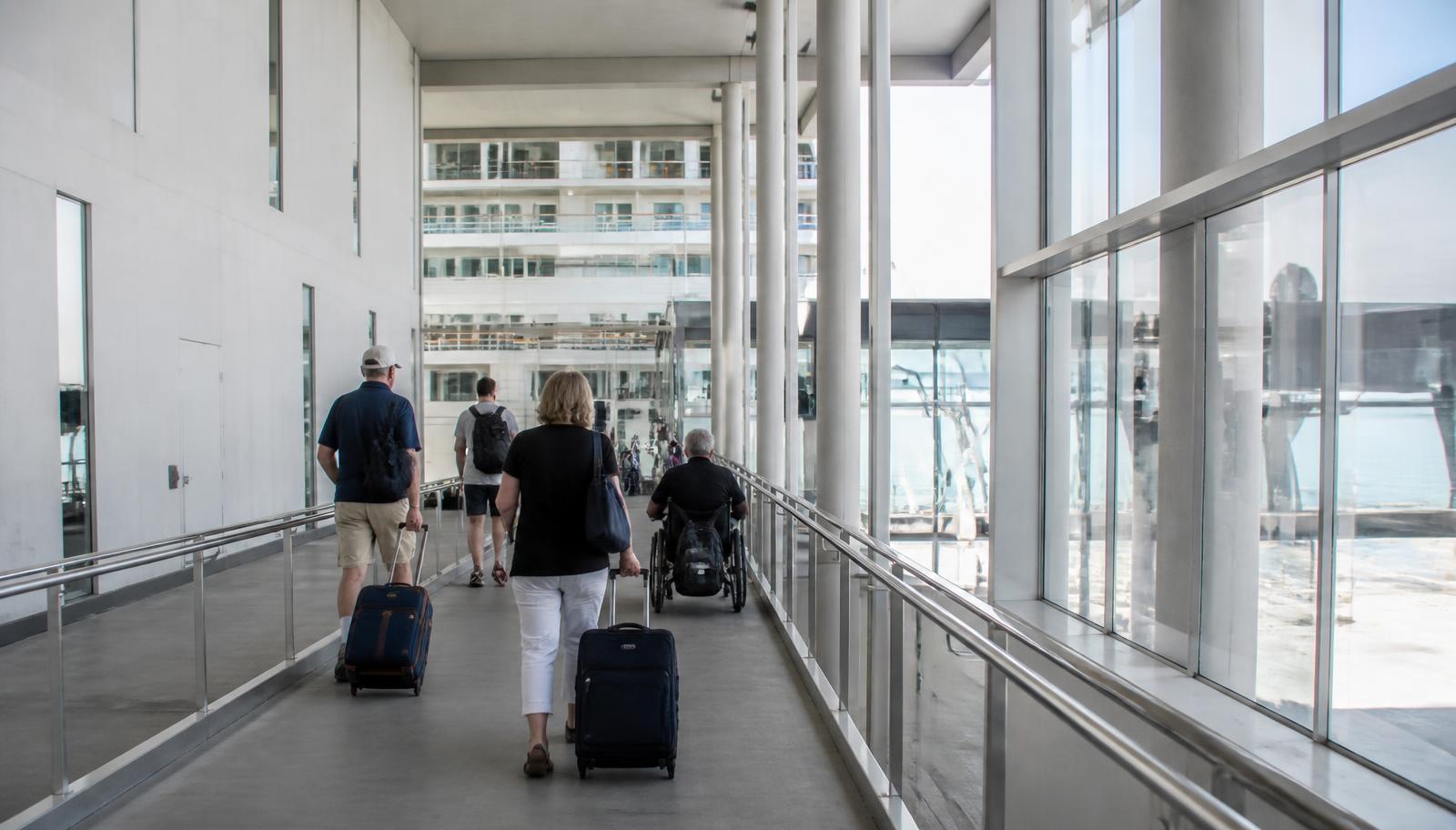 Travellers with wheeled luggage moving along a wide terminal walkway towards a ship, one using a wheelchair