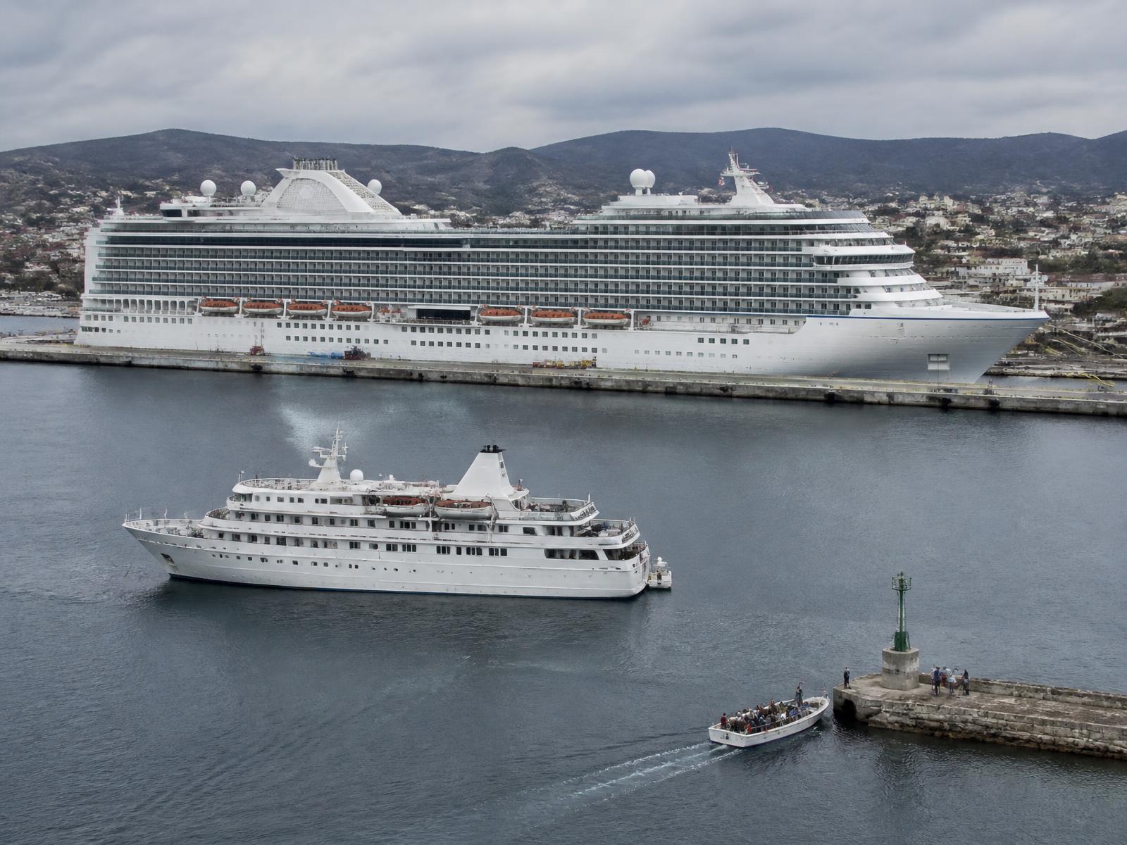 A very large cruise ship alongside a long quay while a much smaller vessel lies at anchor offshore and a tender carries passengers to a stone jetty