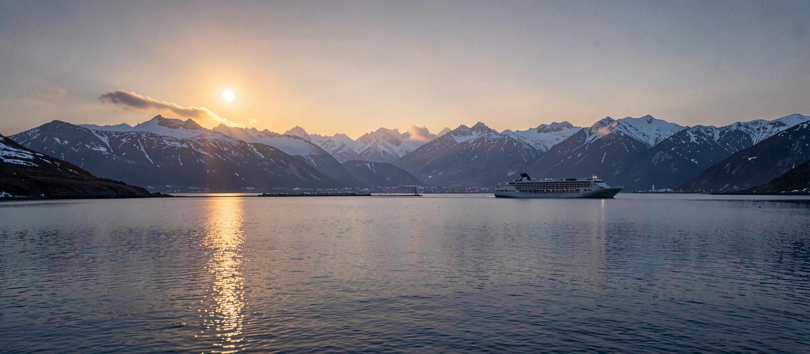Low midnight sun over calm water and snow-streaked mountains at high latitude, with a small passenger ship in the distance